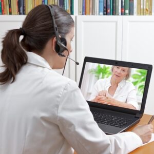 Doctor sitting at the desk of her office with headset and laptop, taking notes during an onlinecall with a female patient showing a large mole, telemedicine concept