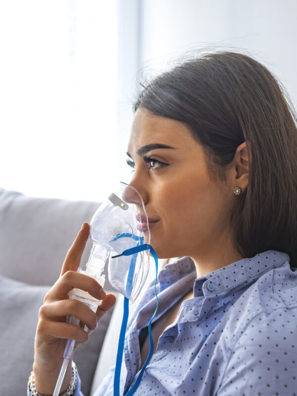 Use nebulizer and inhaler for the treatment. Young woman inhaling through inhaler mask lying on the couch. Woman using asthma machine at home